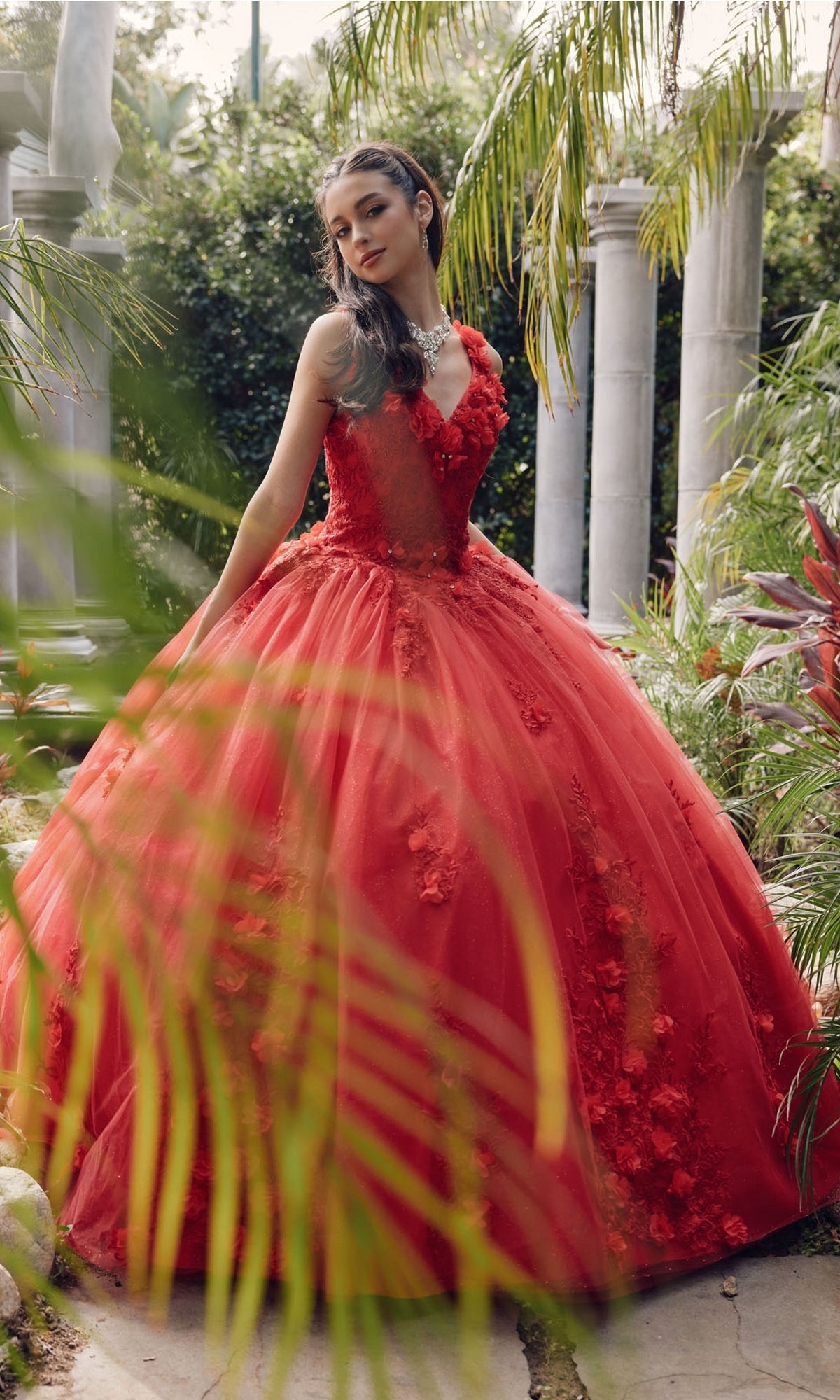 A young woman stands in a garden near stone columns, wearing the Juliet 1437R Red Ball Gown Quinceañera Dress with floral details and beaded embroidery, surrounded by lush tropical plants.